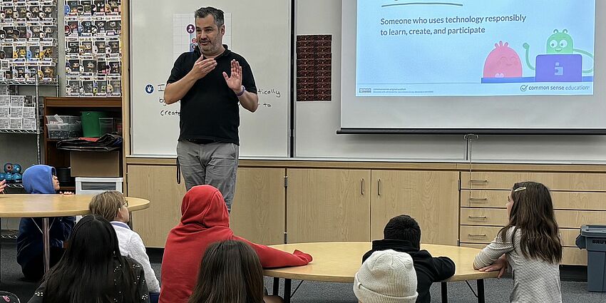 male teacher stands at white board that has the words Digital Citizen as several students sit on the floor listening to him