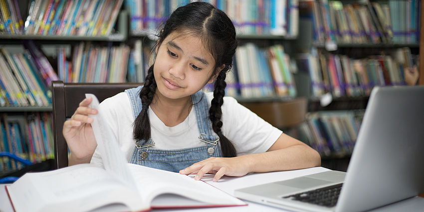 adolescent girl sitting at a table in a library is looking at a book and has a laptop computer open in front of her