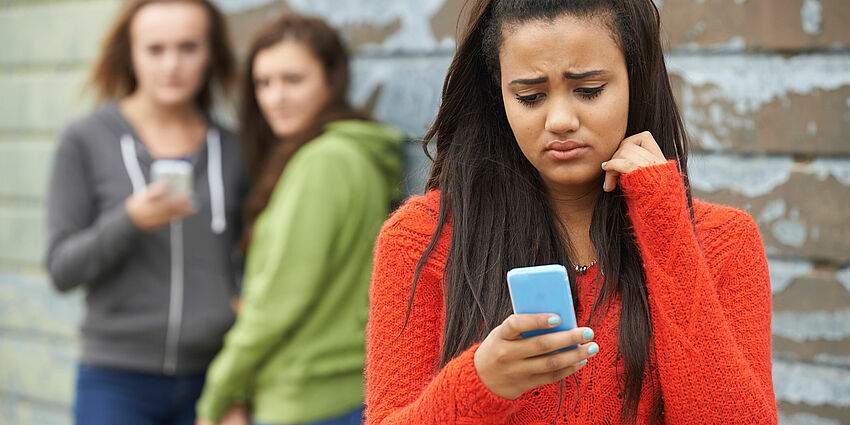 sad teen in foreground looking at cell phone as 2 girls in background look on with sneers.