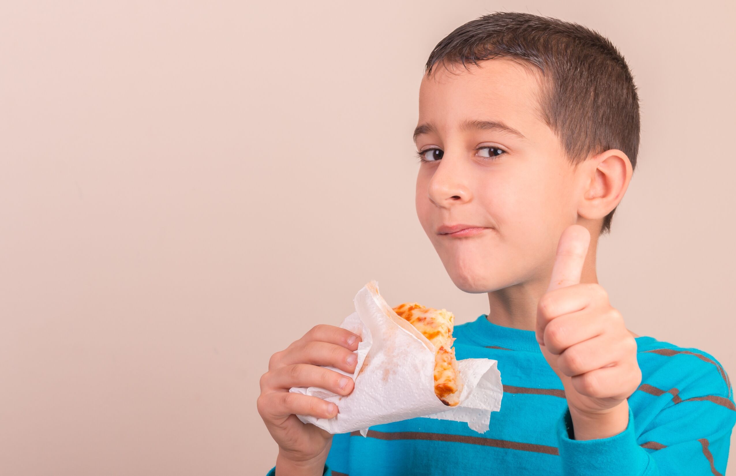 boy holding a slice of pizza in one hand and giving a Thumbs Up with the other.